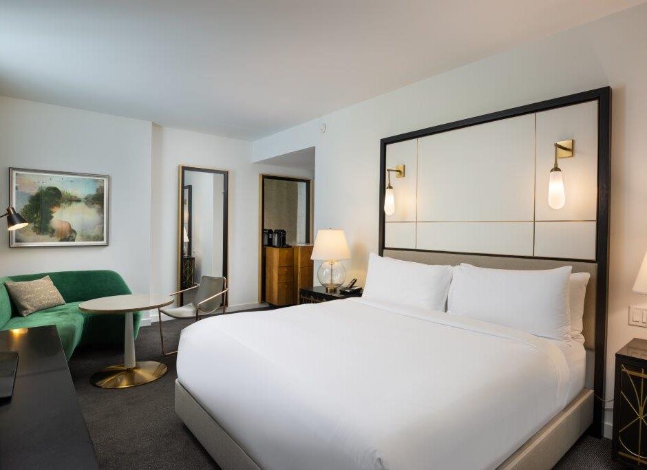 Modern hotel room with a king bed, green loveseat, round table, wall art, lamps, and a mirror, decorated in neutral and gold tones.