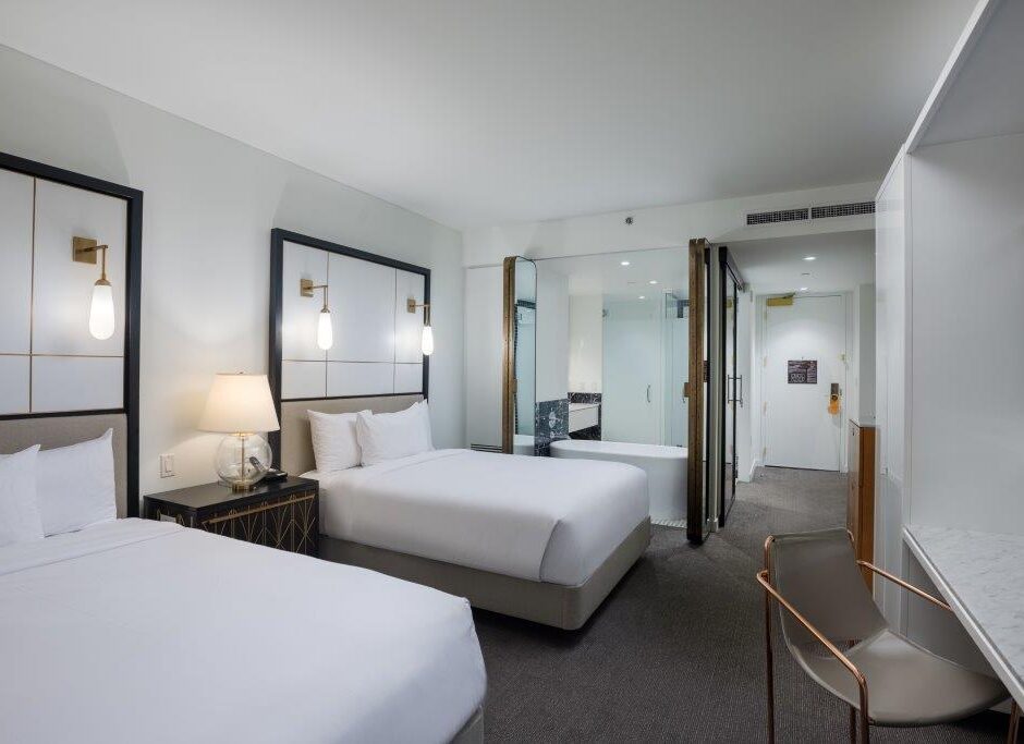 Hotel room with two double beds, a nightstand, lamps, desk, chair, TV, and a large mirror. The bathroom is visible in the background.