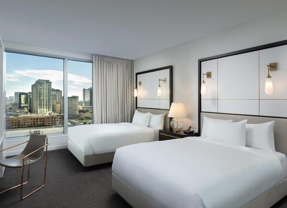 Modern hotel room with two double beds, a desk and chair, large window, city skyline view, neutral decor, and wall-mounted lights above the beds.