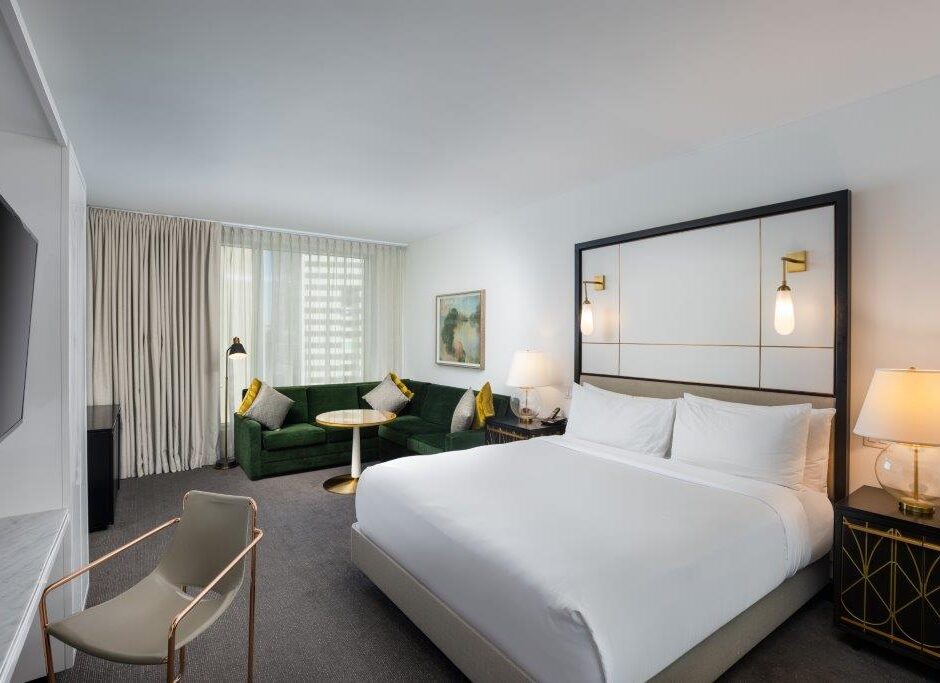 Modern hotel room with a king bed, green sofa, gold accents, large window with curtains, and a wall-mounted TV.