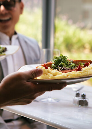 A person hands a plate of mashed potatoes and vegetables to someone at a restaurant table, with another diner smiling in the background.