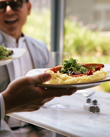 A person hands a plate of mashed potatoes and vegetables to someone at a restaurant table, with another diner smiling in the background.