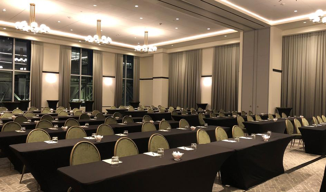 A large conference room with rows of tables and chairs, each set with glasses, notepads, and pens, under bright ceiling lights.