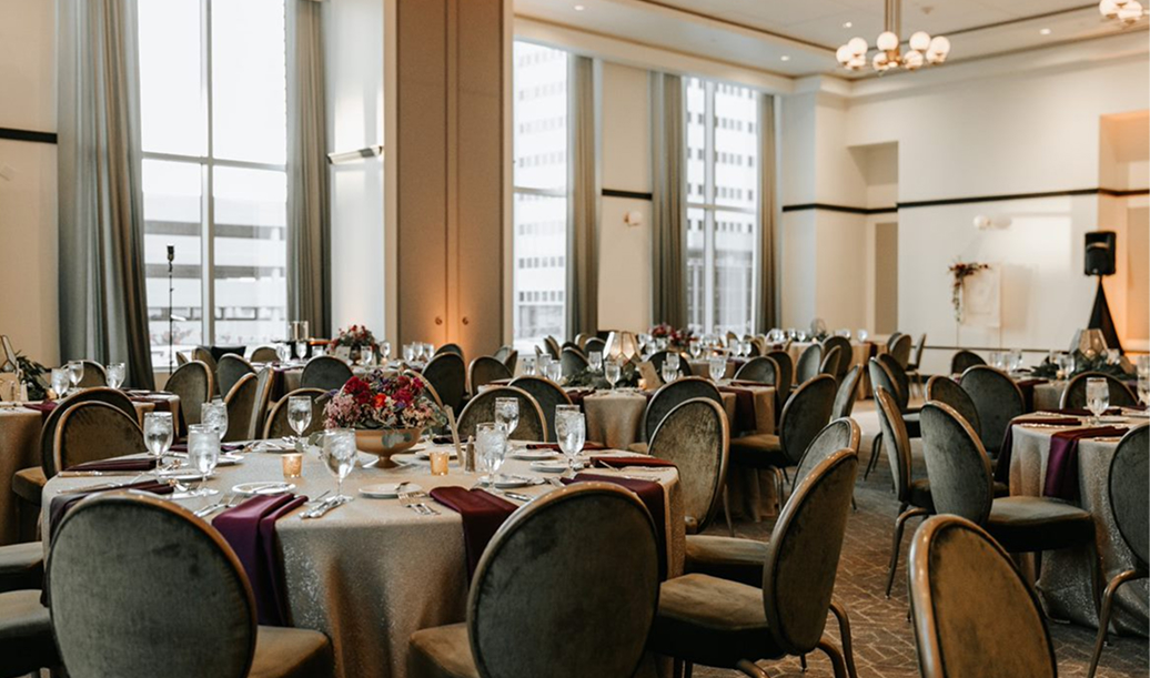 Elegant banquet hall with round tables set for an event, featuring floral centerpieces, glassware, and floor-to-ceiling windows.