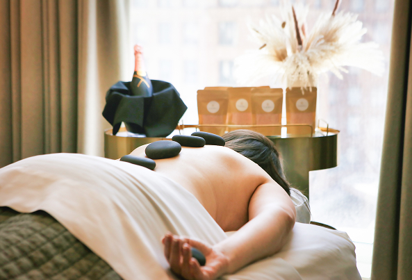 Person lying face down during a hot stone massage, with stones on their back, near a window with a champagne bottle and flowers on a table.
