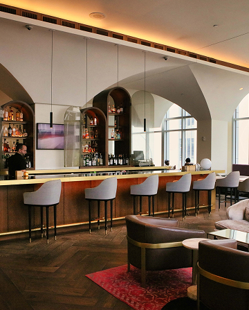 Modern bar with high stools, shelves of liquor, a bartender, and arched mirrors. Soft lighting and contemporary decor create a cozy atmosphere.