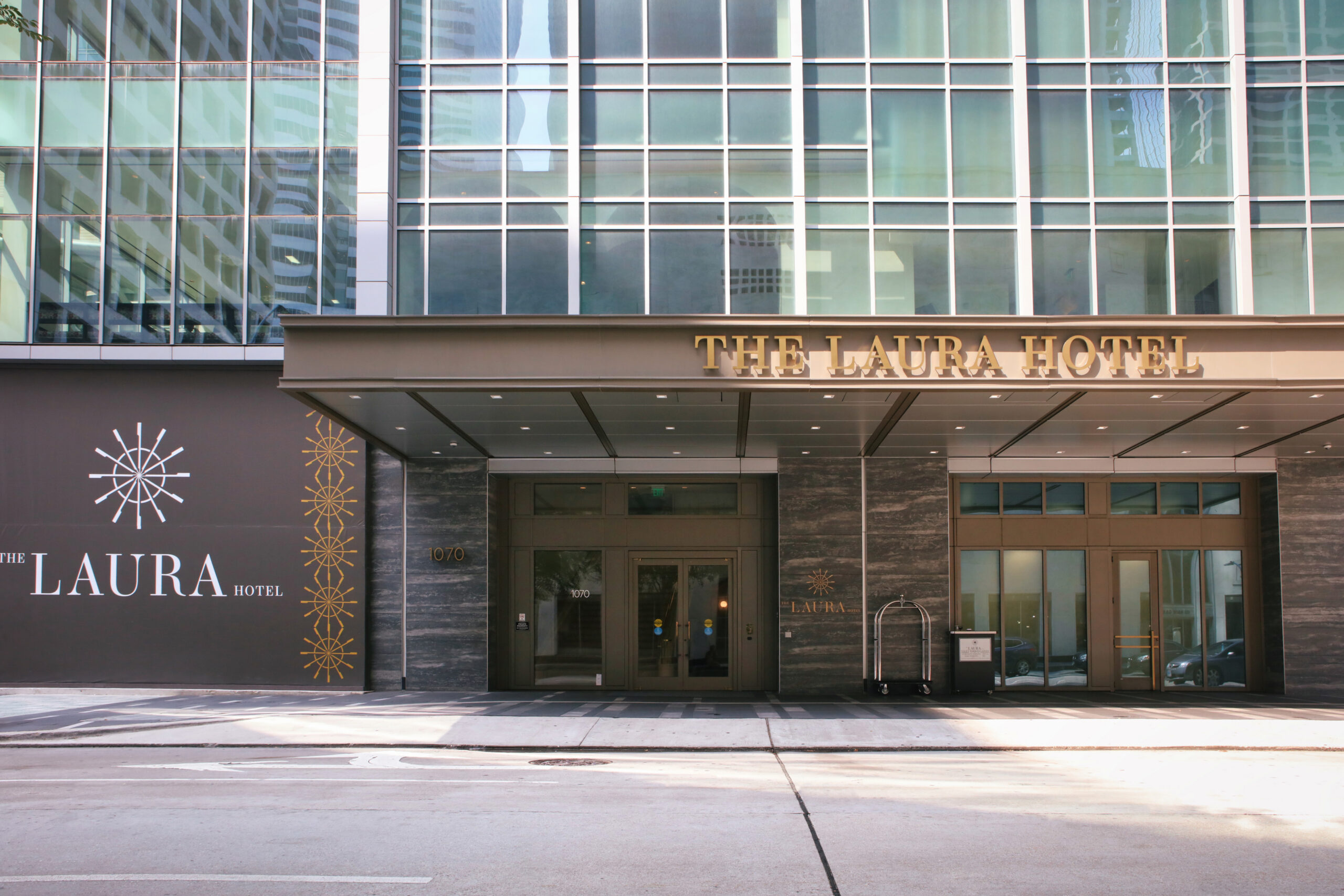 The exterior entrance of The Laura Hotel features glass doors, tall windows, and a sign with the hotels name above the entrance.