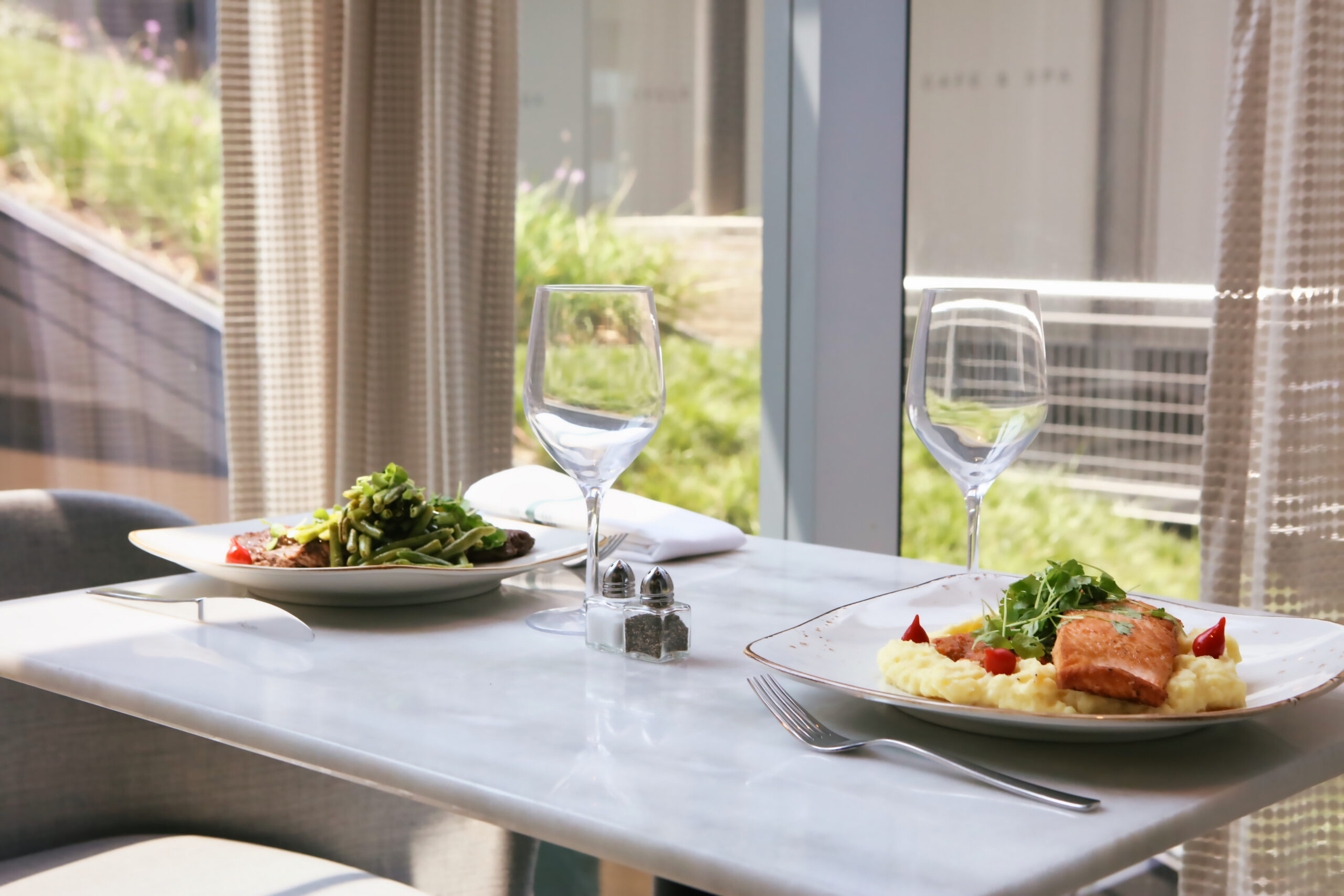 A table for two with plates of food, empty wine glasses, and utensils by a window overlooking greenery.