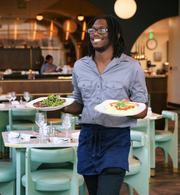 A smiling server wearing glasses and a blue apron carries two plates of food in a modern, stylish restaurant with green chairs and set tables.