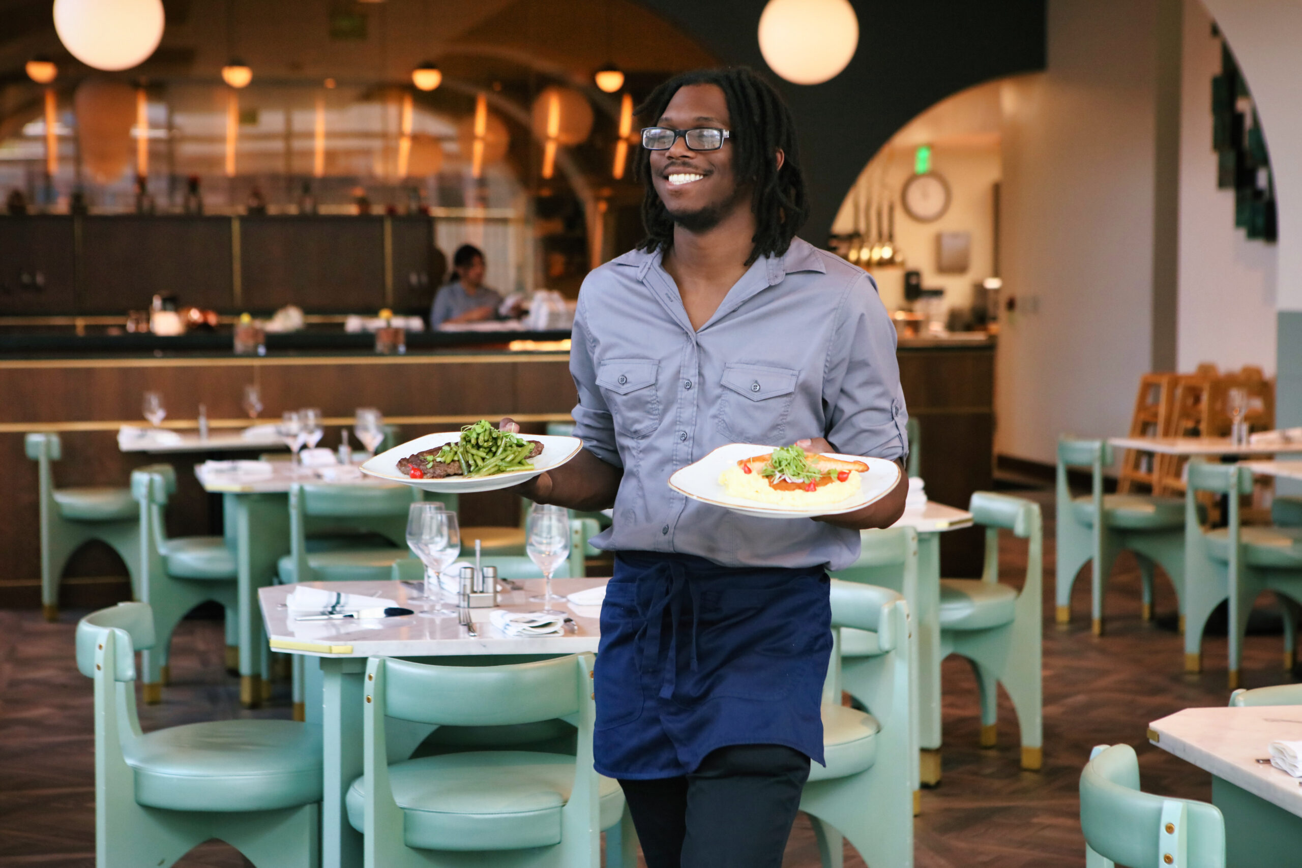 A smiling server wearing glasses and a blue apron carries two plates of food in a modern, stylish restaurant with green chairs and set tables.
