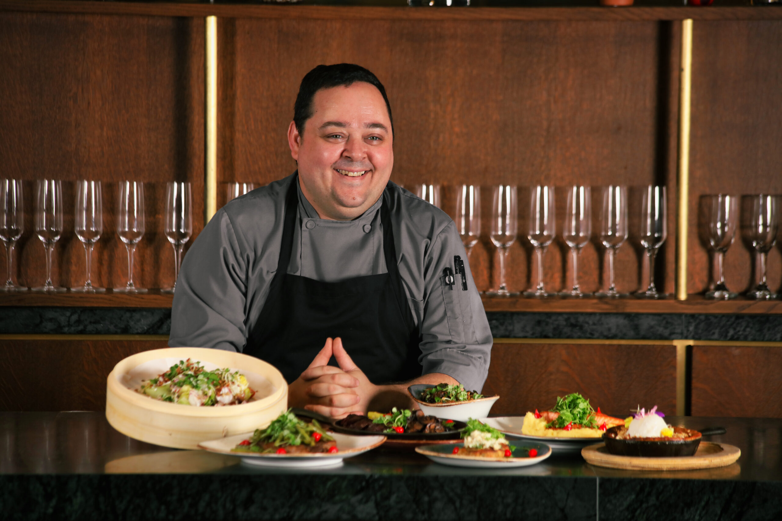 Smiling chef in a gray uniform stands behind a counter with several plated dishes and empty wine glasses lined up on shelves behind him.