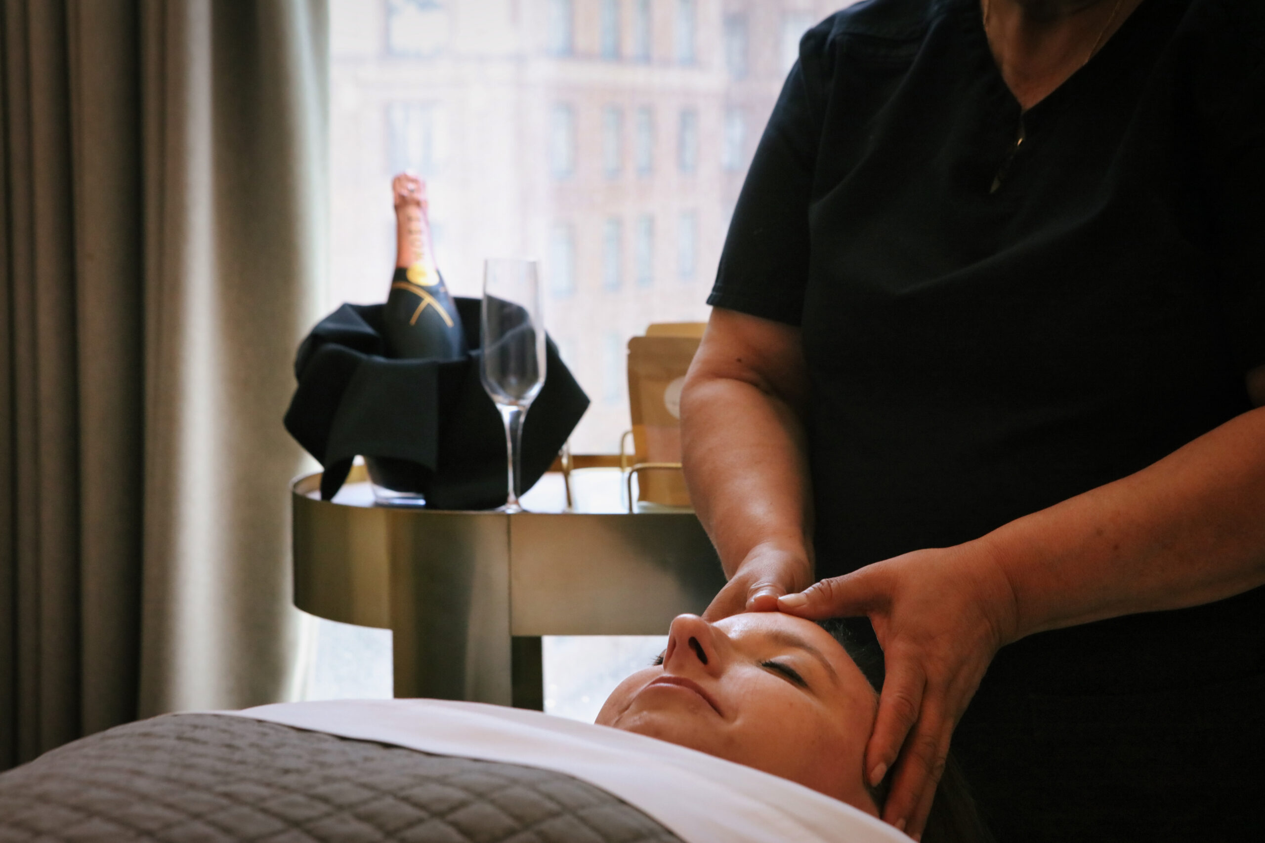 A person receives a facial massage in a spa, with a bottle of champagne and an empty glass on a table nearby.