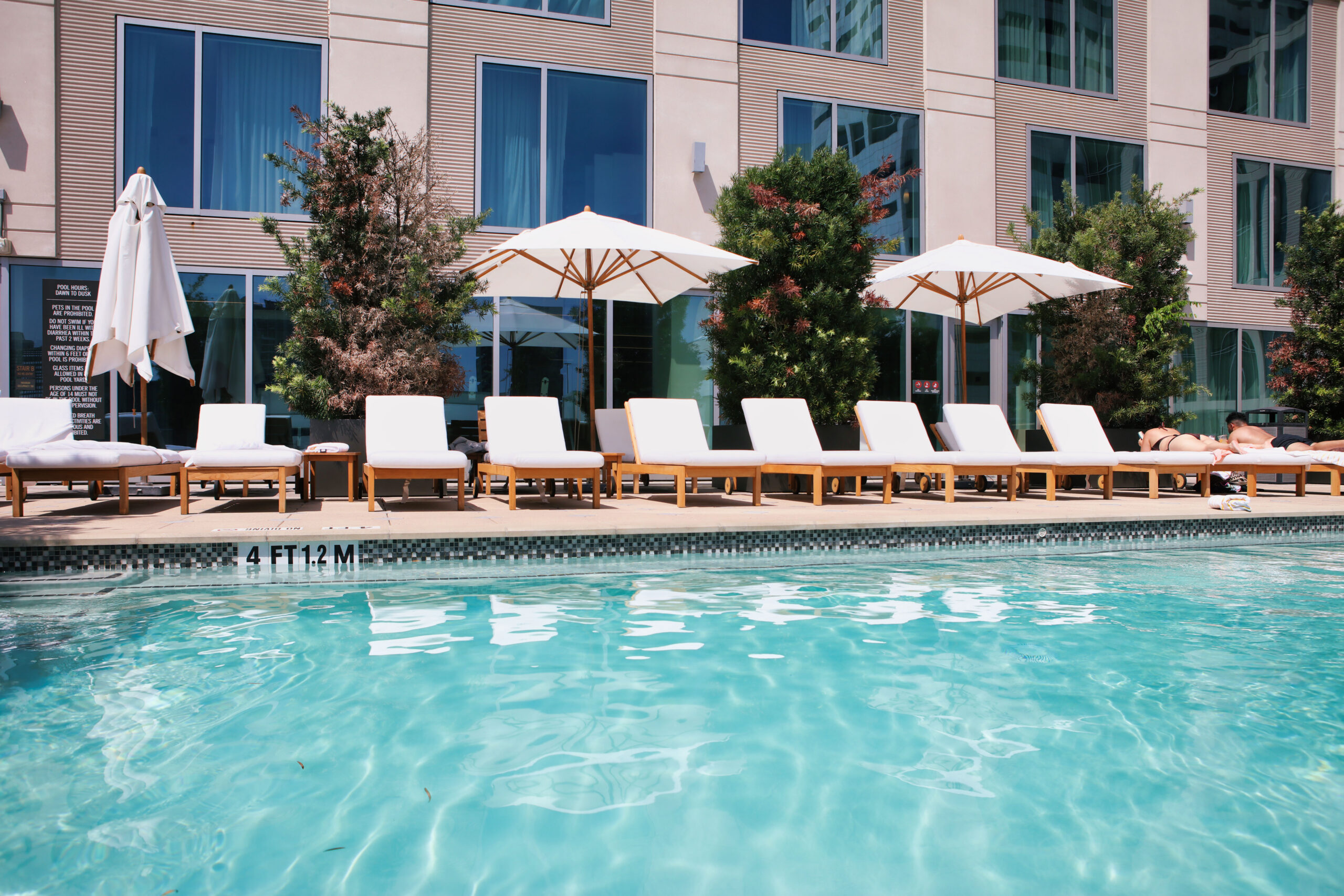 Outdoor pool with clear water, lined with white lounge chairs and umbrellas, near a modern building with large windows and some greenery.