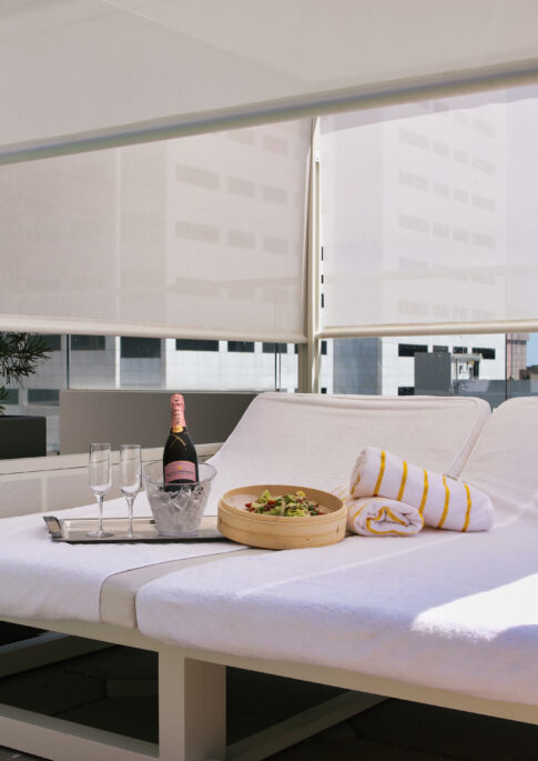 Two lounge chairs with white towels, a tray with champagne and glasses, and a bowl of salad under a cabana on a sunny rooftop.