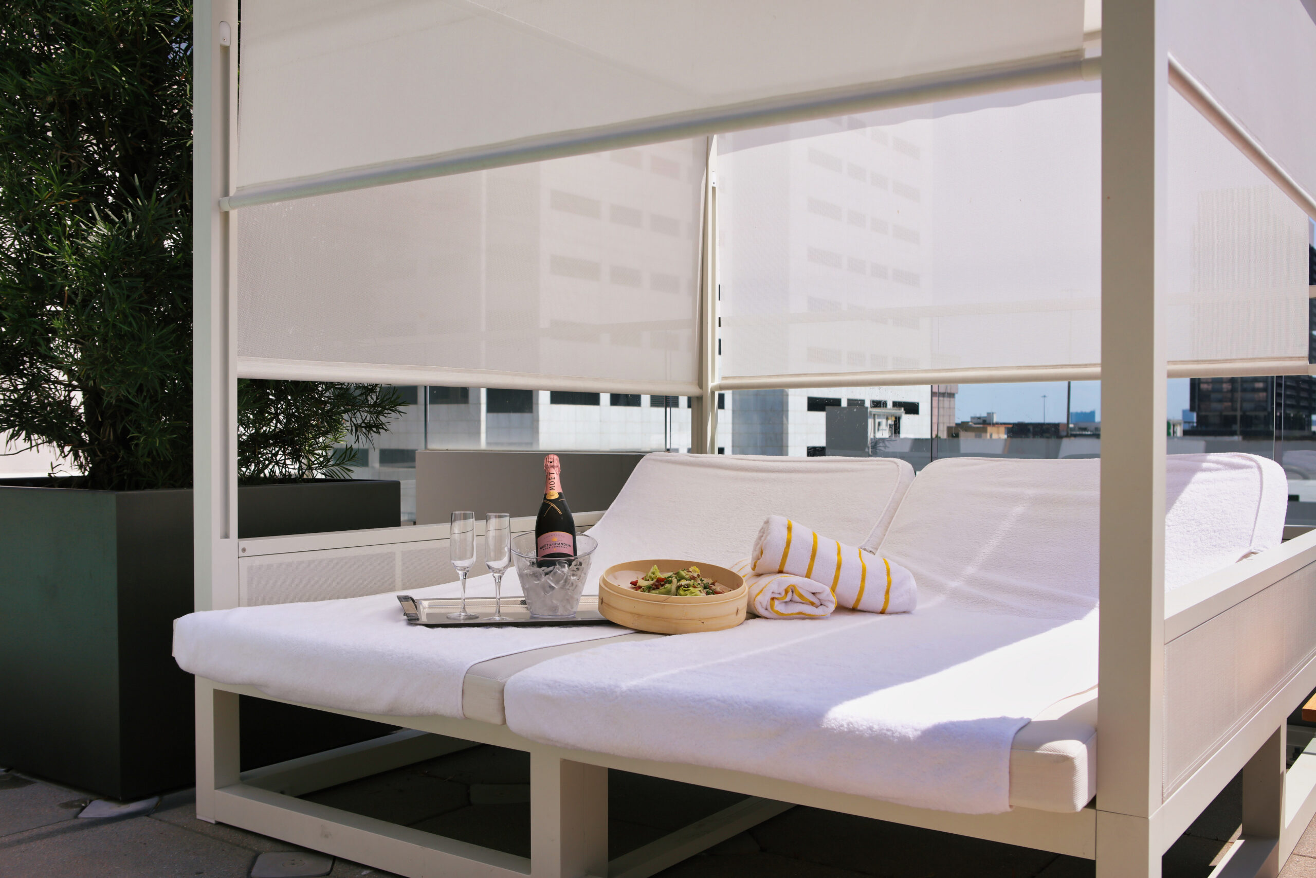 Two lounge chairs with white towels, a tray with champagne and glasses, and a bowl of salad under a cabana on a sunny rooftop.