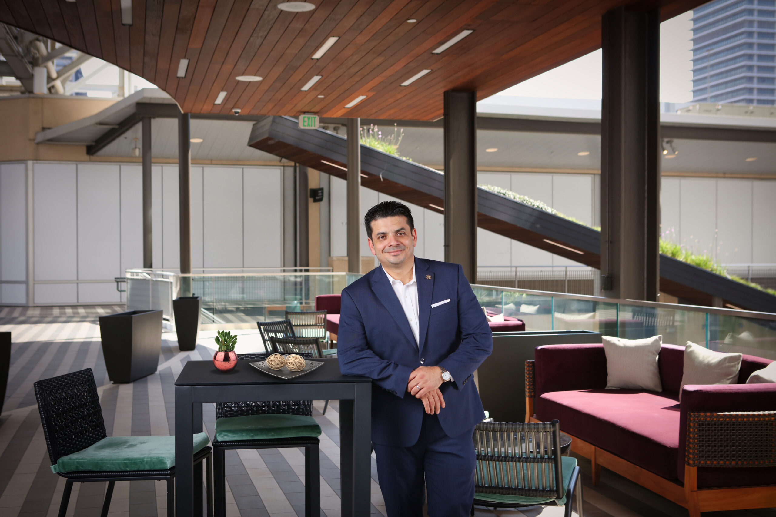 A man in a blue suit leans on a black table in a modern outdoor lounge with chairs, couches, and wooden ceiling overhead.