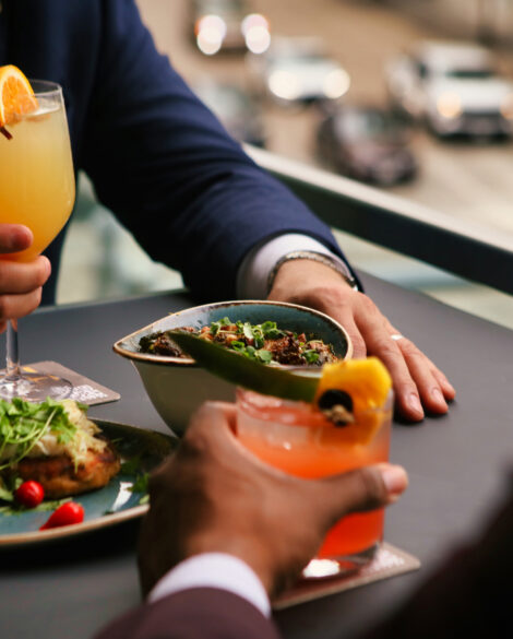 Two people in suits enjoying cocktails and plates of food at an outdoor restaurant table overlooking a city street.