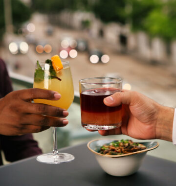 Two people clink cocktails at an outdoor table, with a bowl of snacks and a blurred city street in the background.