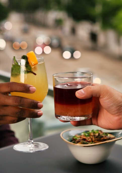 Two people clink cocktails at an outdoor table, with a bowl of snacks and a blurred city street in the background.