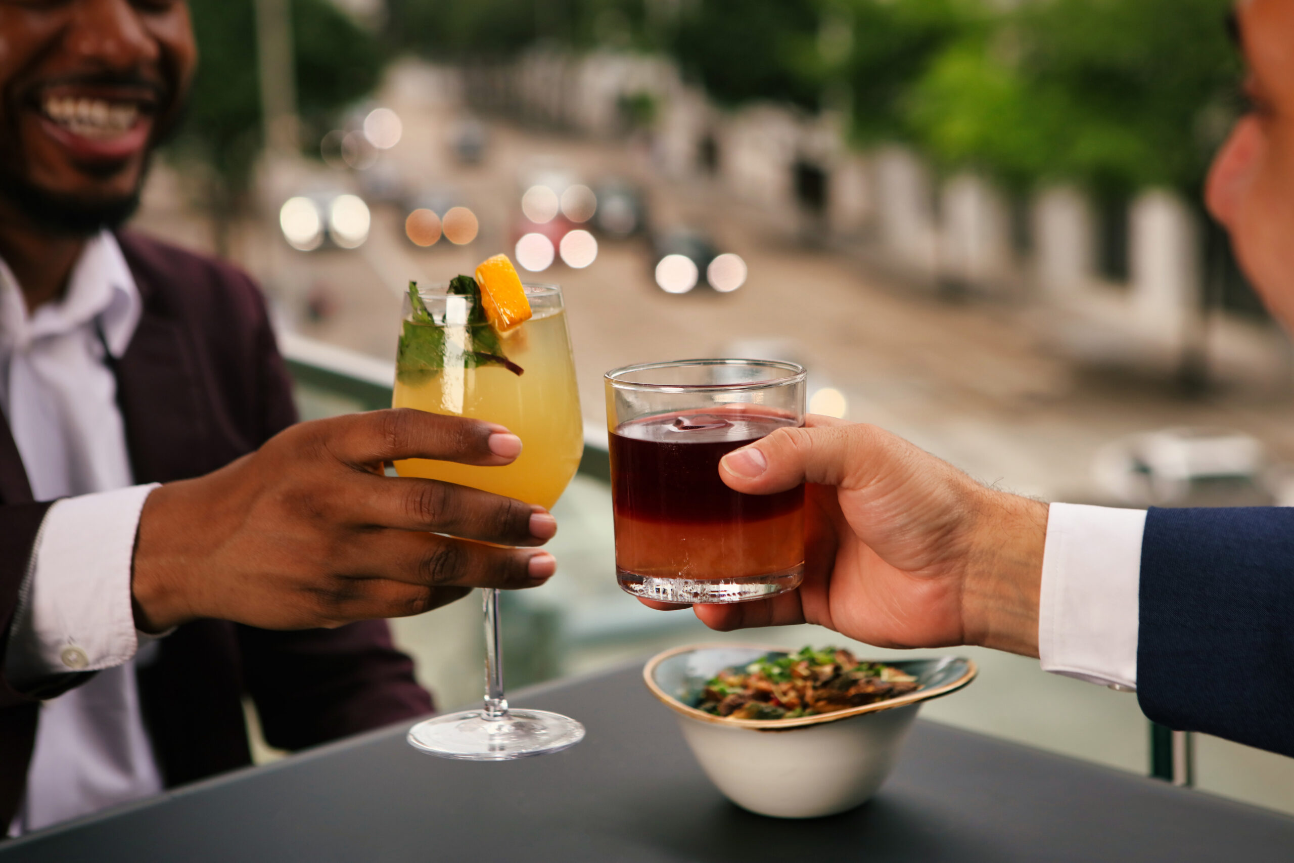 Two people clink cocktails at an outdoor table, with a bowl of snacks and a blurred city street in the background.