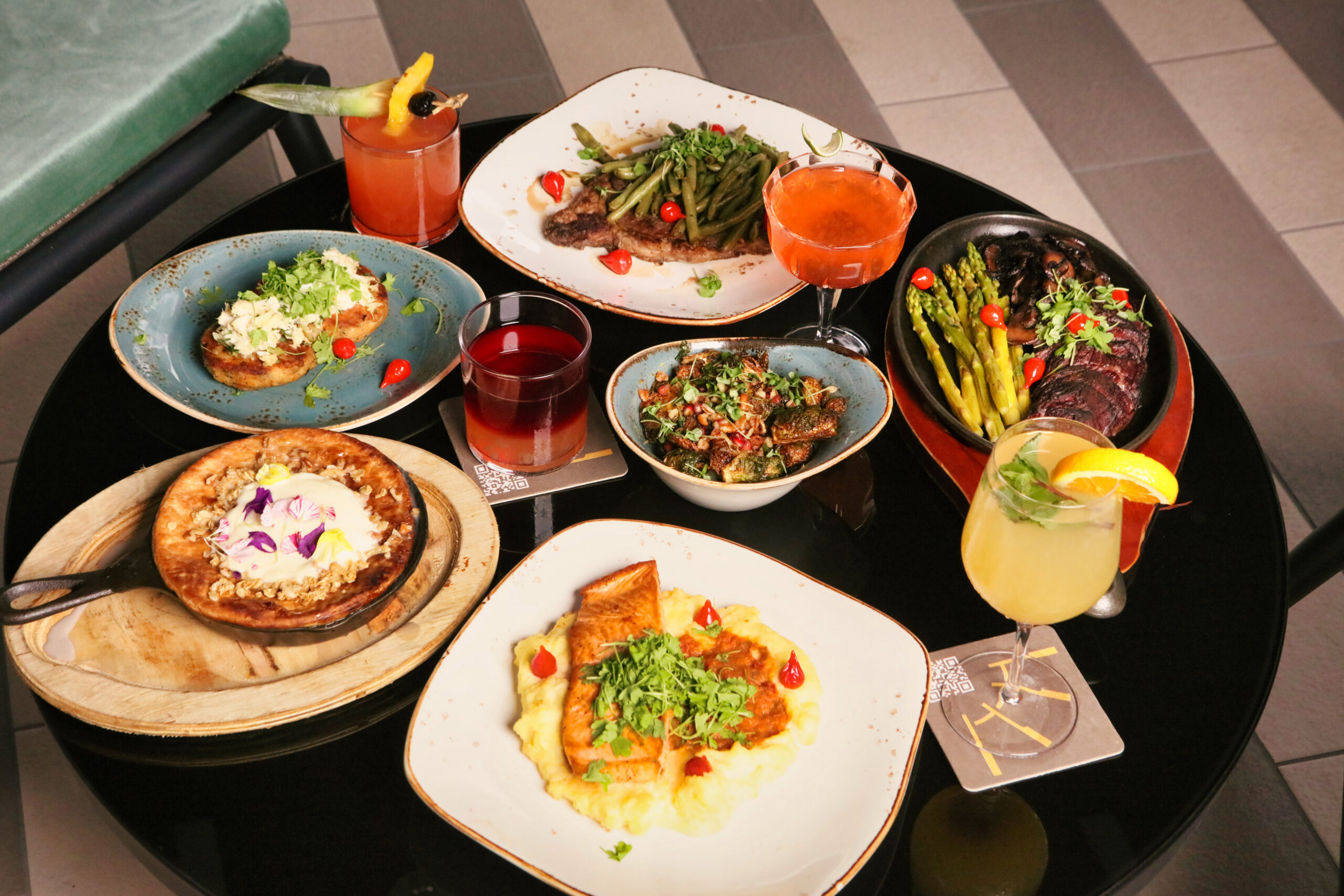 A round table with assorted dishes, including steak, vegetables, drinks, toast, and salad, arranged on colorful plates and glasses.