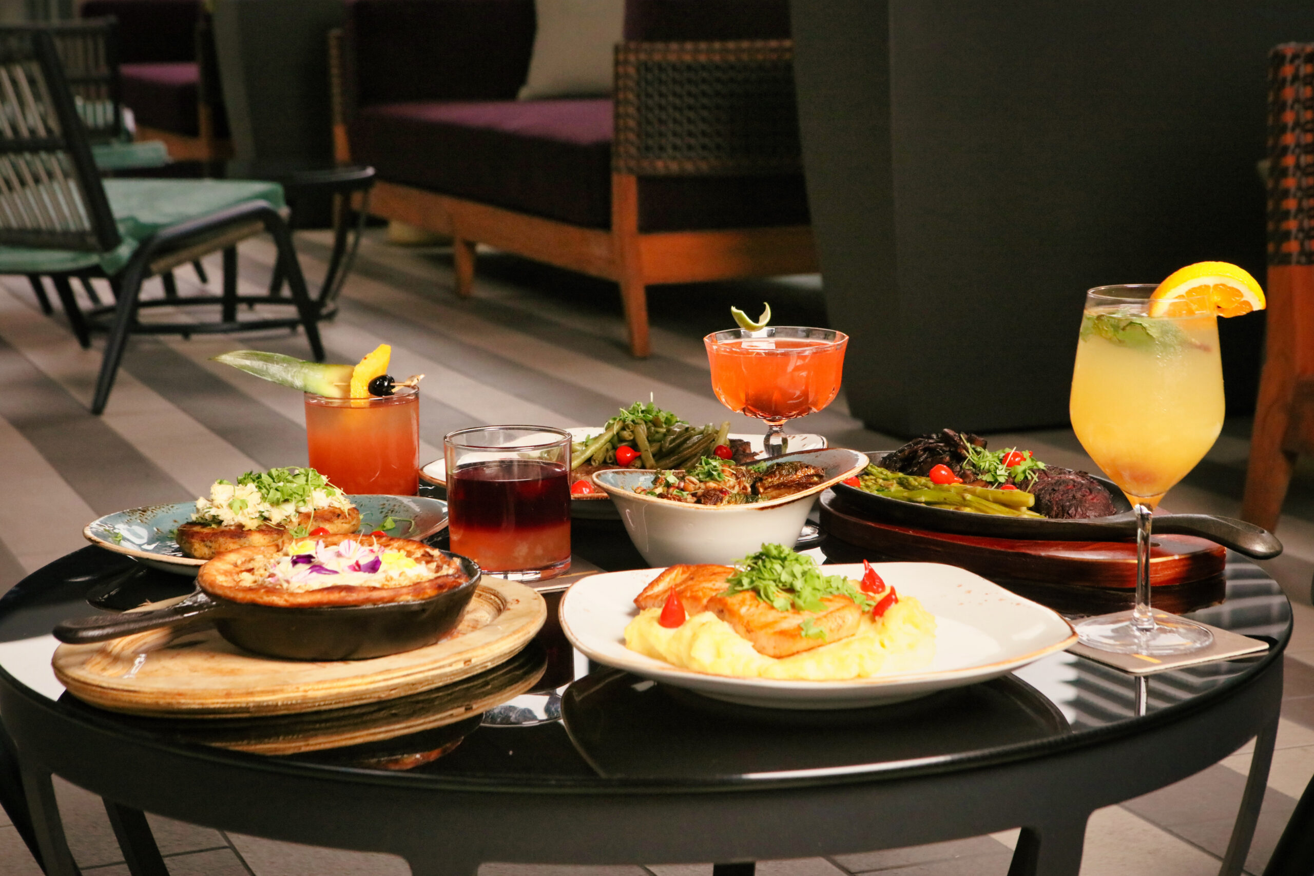 A round table with assorted dishes, cocktails, and desserts, set in an indoor lounge with chairs in the background.