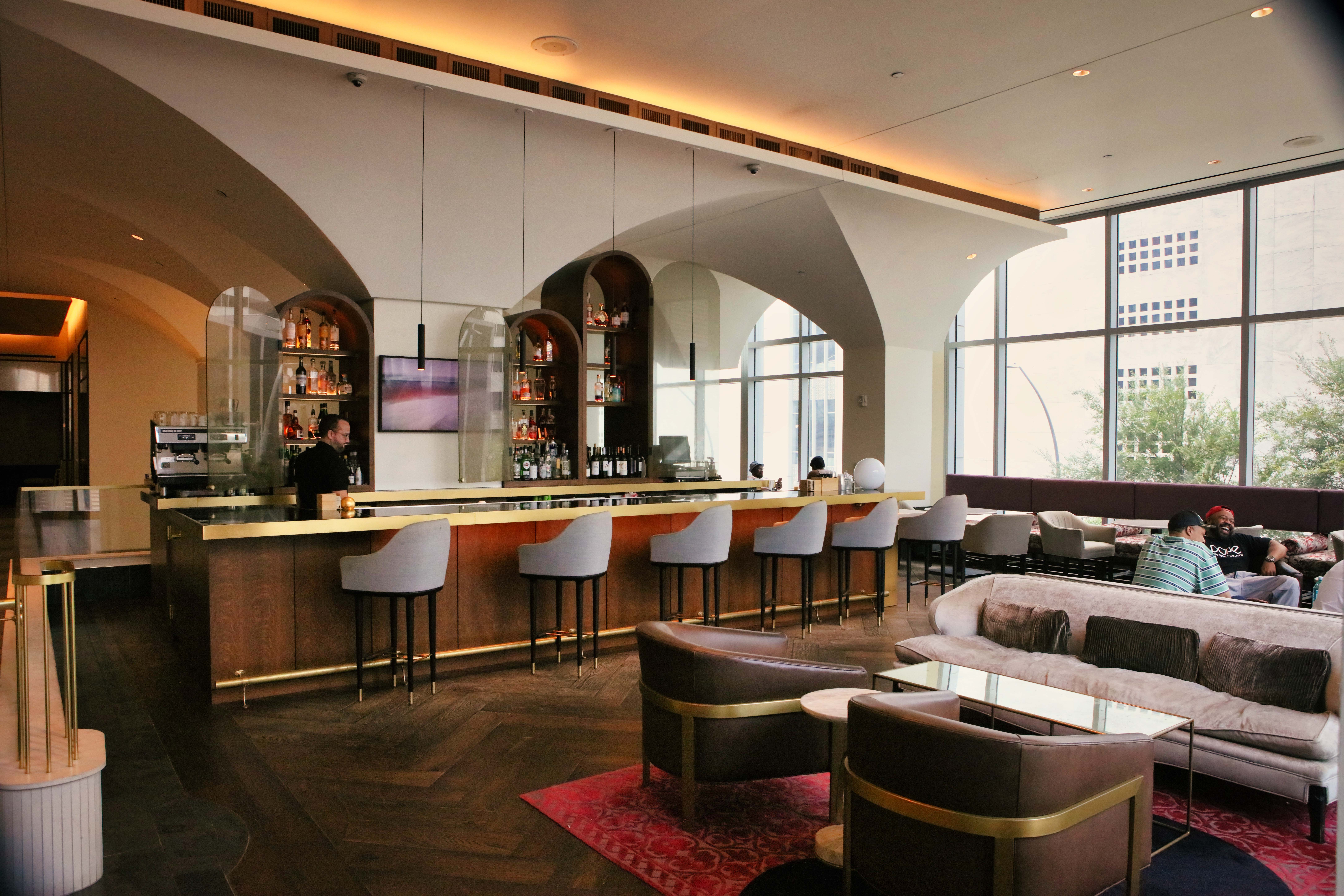 Modern hotel bar with high chairs, a bartender, shelves of bottles, and guests sitting on sofas near large windows with city views.