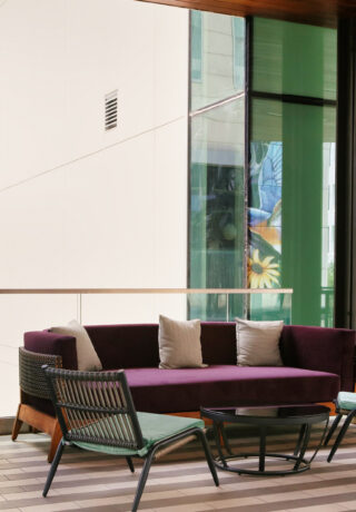 Outdoor lounge with a purple sofa, two chairs, a glass table, and a TV mounted above, on a balcony with tall glass railing and city view.