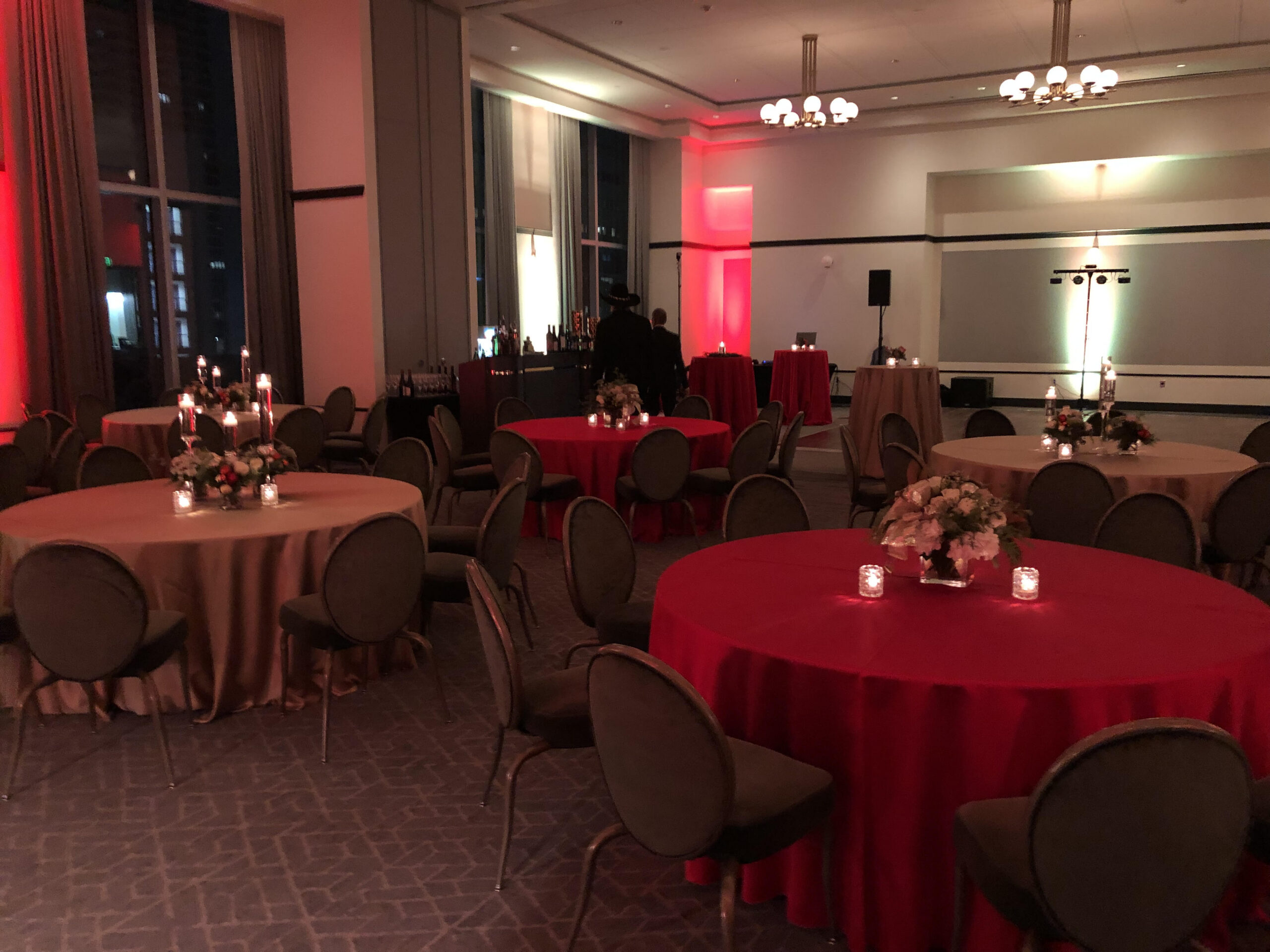 Elegant banquet hall with round tables, red and beige tablecloths, floral centerpieces, candles, and soft red lighting.