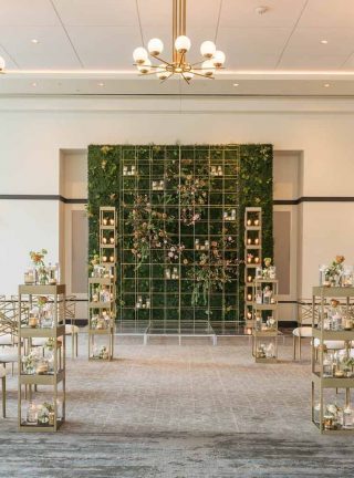 Elegant wedding ceremony setup with gold chairs, small flower arrangements, and a green wall backdrop with candles in a bright, modern room.