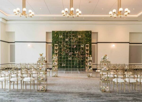 Elegant wedding ceremony setup with gold chairs, small flower arrangements, and a green wall backdrop with candles in a bright, modern room.