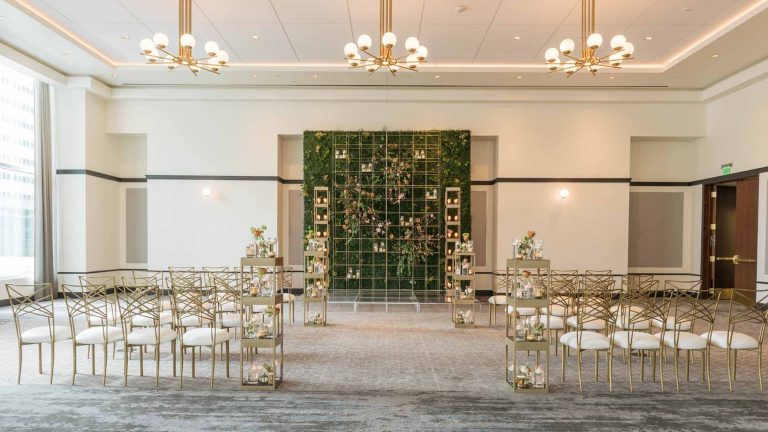Elegant wedding ceremony setup with gold chairs, small flower arrangements, and a green wall backdrop with candles in a bright, modern room.