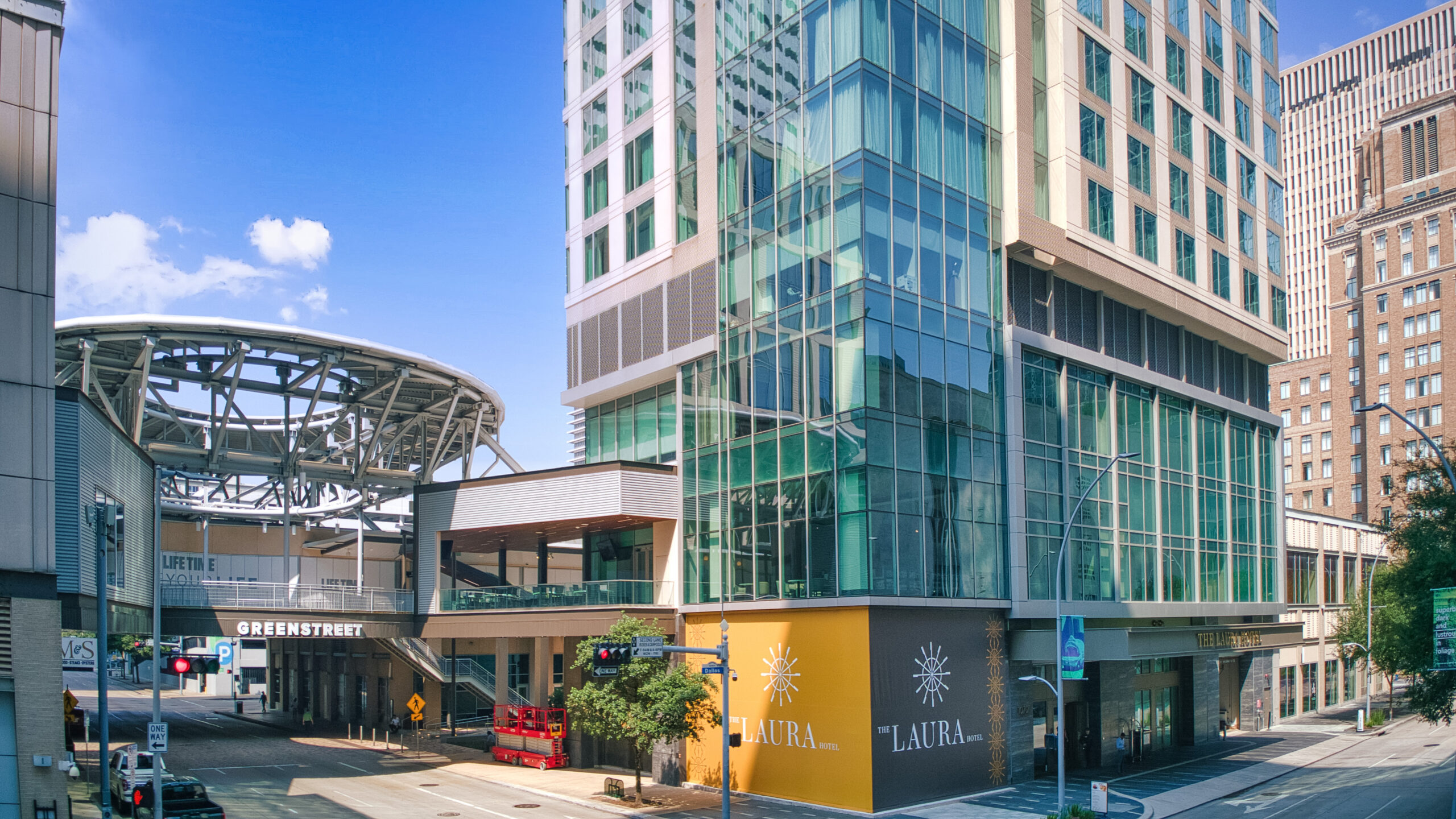 Modern city street corner with tall glass buildings, GreenStreet sign, yellow storefronts, and a circular metal structure under a blue sky.