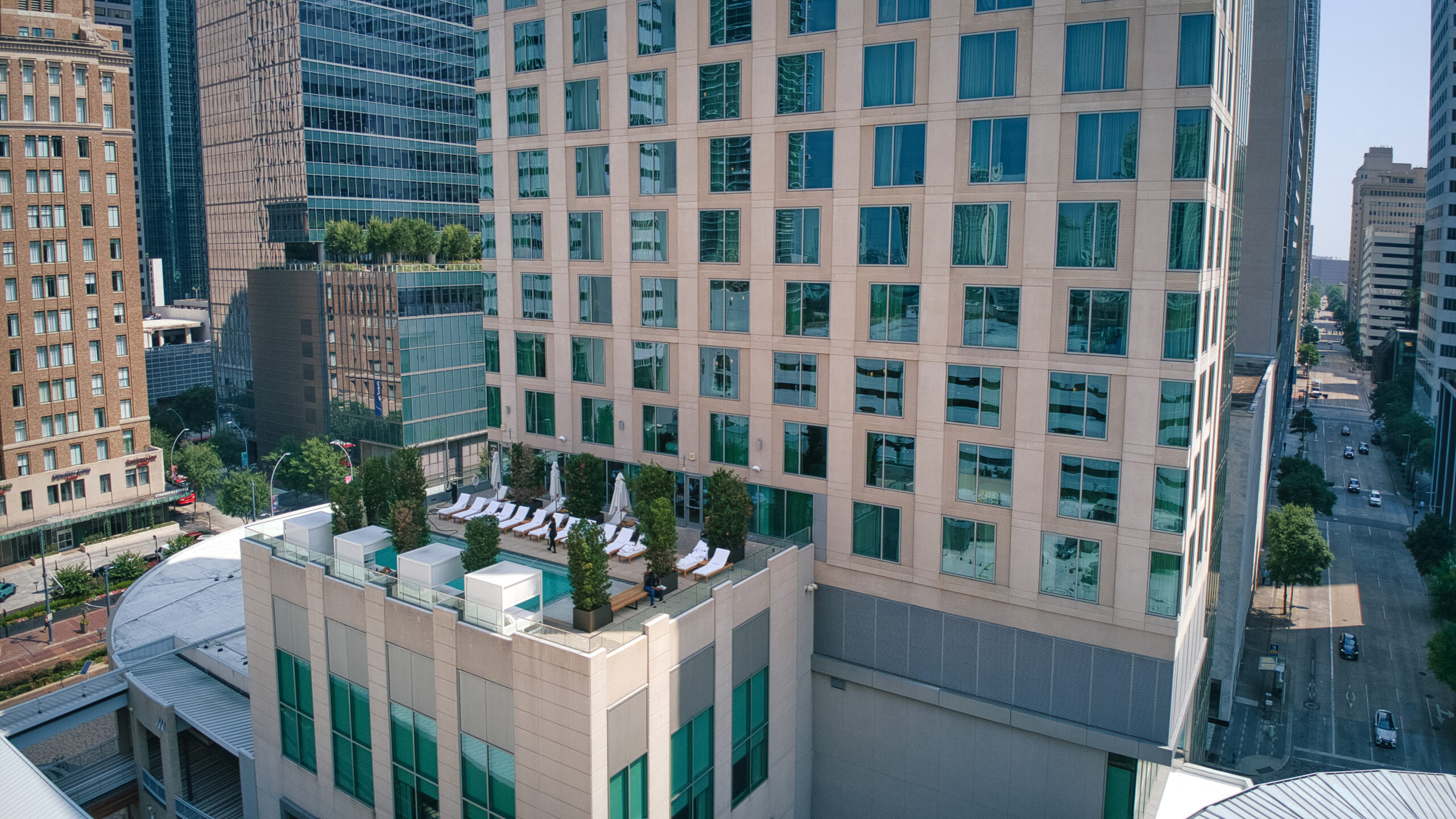 A rooftop pool with lounge chairs and umbrellas sits atop a tall city building, surrounded by other modern skyscrapers and urban streets.