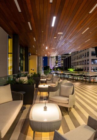 Night terrace with cozy seating, wooden ceiling, and a hummingbird mural, overlooking a city street and surrounding buildings.