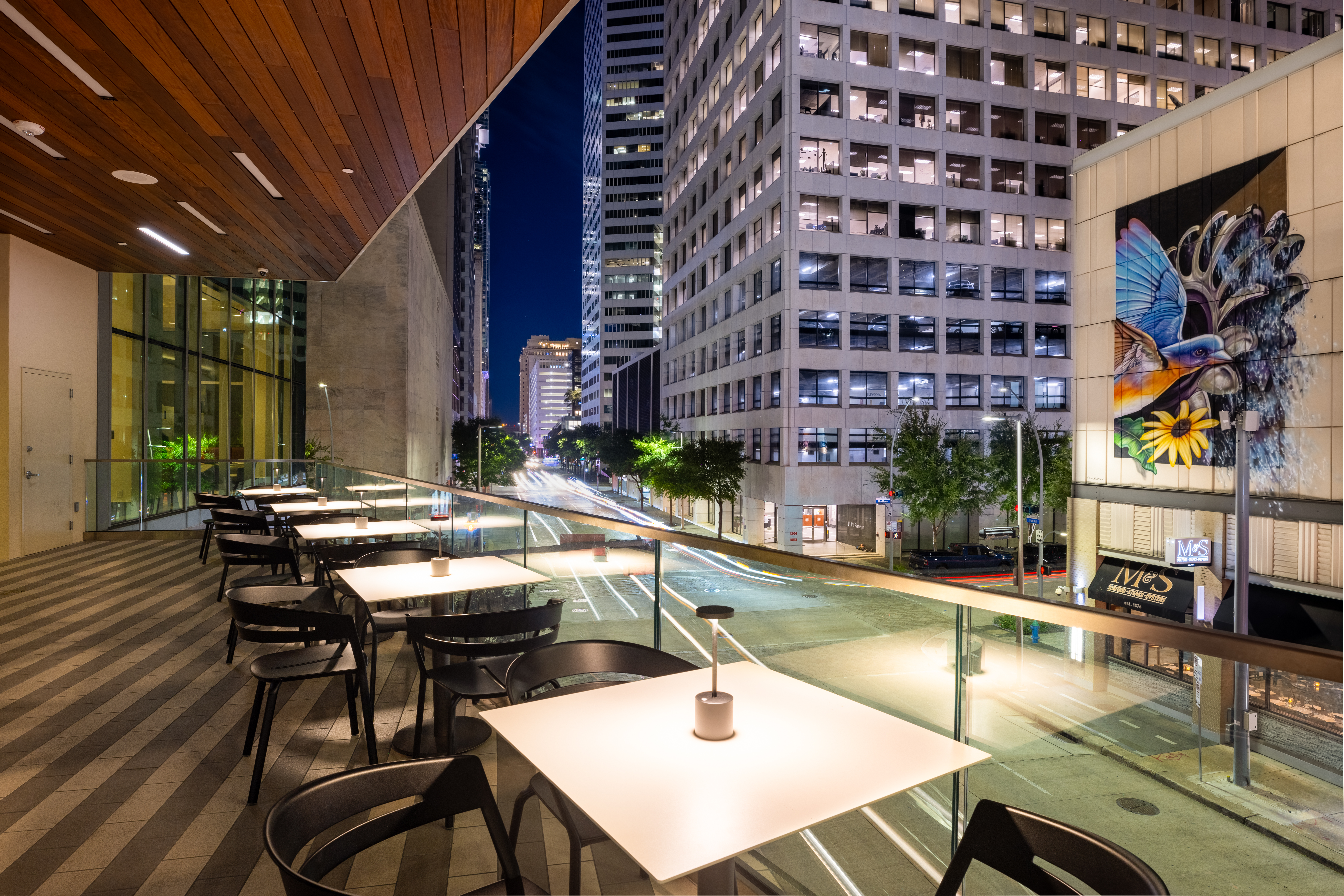 Outdoor hotel terrace with dining tables overlooking downtown Houston streets, high-rise buildings and a colorful bird mural at night
