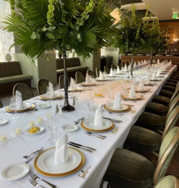 Long banquet table with white tablecloth, gold-rimmed plates, folded napkins, and tall green floral centerpieces set for a formal event.