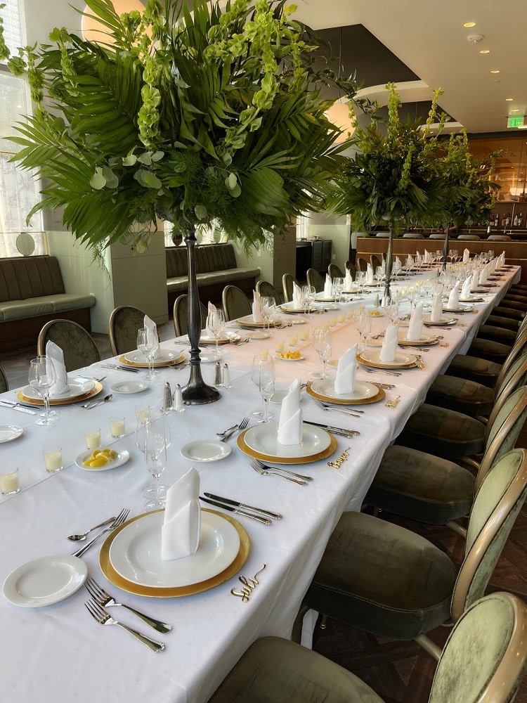 Long banquet table with white tablecloth, gold-rimmed plates, folded napkins, and tall green floral centerpieces set for a formal event.