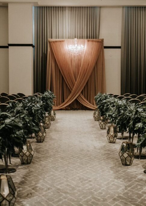 A wedding ceremony setup with rows of chairs, greenery-lined aisle, lanterns, and a peach draped backdrop with a chandelier.