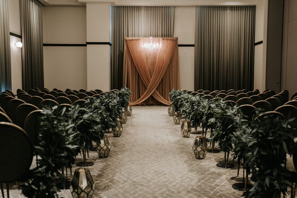 A wedding ceremony setup with rows of chairs, greenery-lined aisle, lanterns, and a peach draped backdrop with a chandelier.