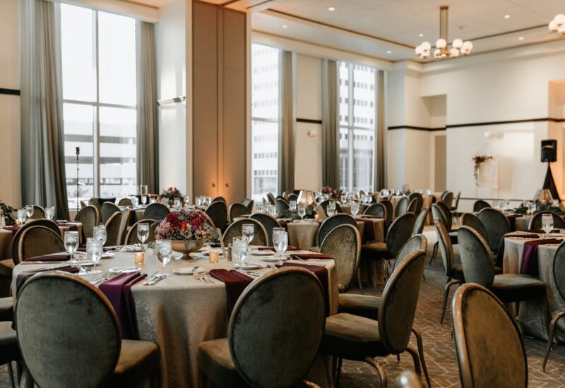 Elegant banquet hall with round tables set for an event, featuring floral centerpieces and large windows letting in natural light.