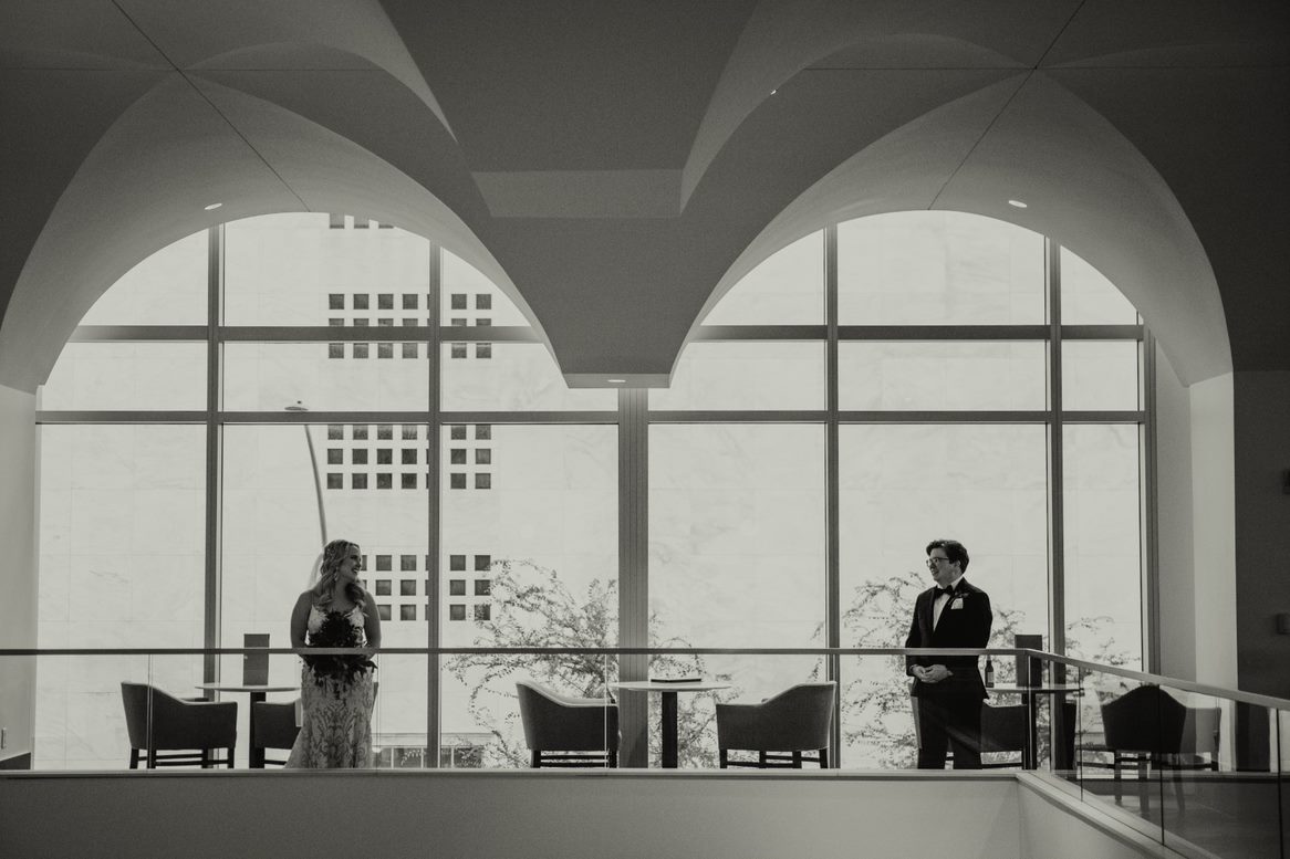 A woman in a gown and a man in a tuxedo stand apart on a balcony, facing each other with large windows in the background.