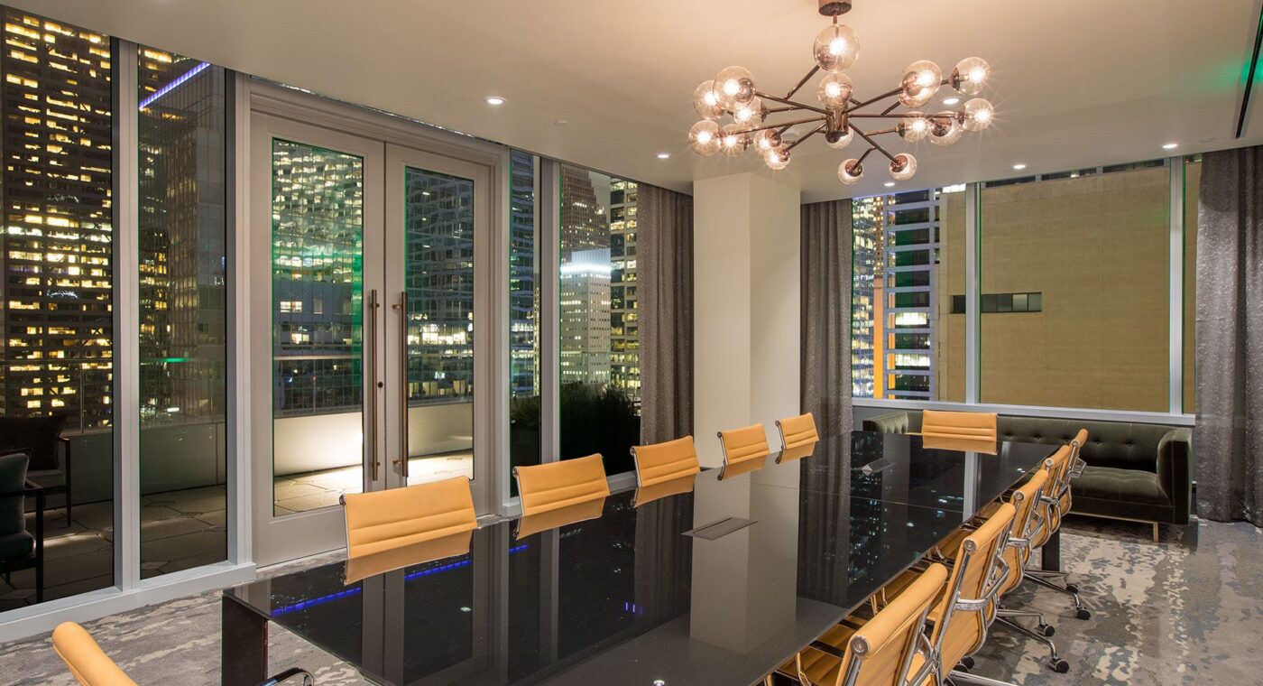 Modern conference room with a long black table, tan chairs, city view through large windows, and a chandelier overhead at night.