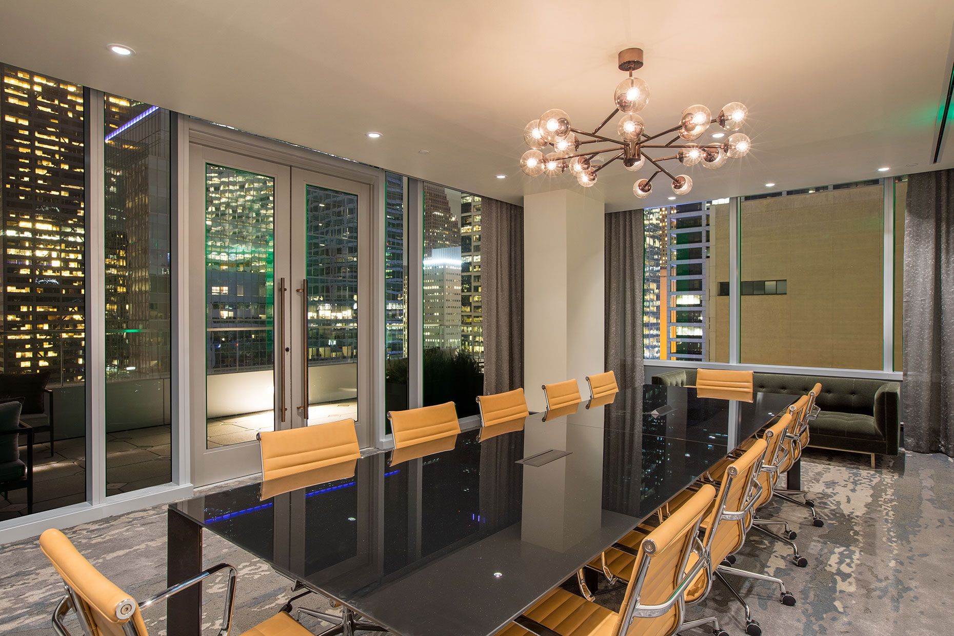Modern conference room with a long black table, tan chairs, city view through large windows, and a chandelier overhead at night.