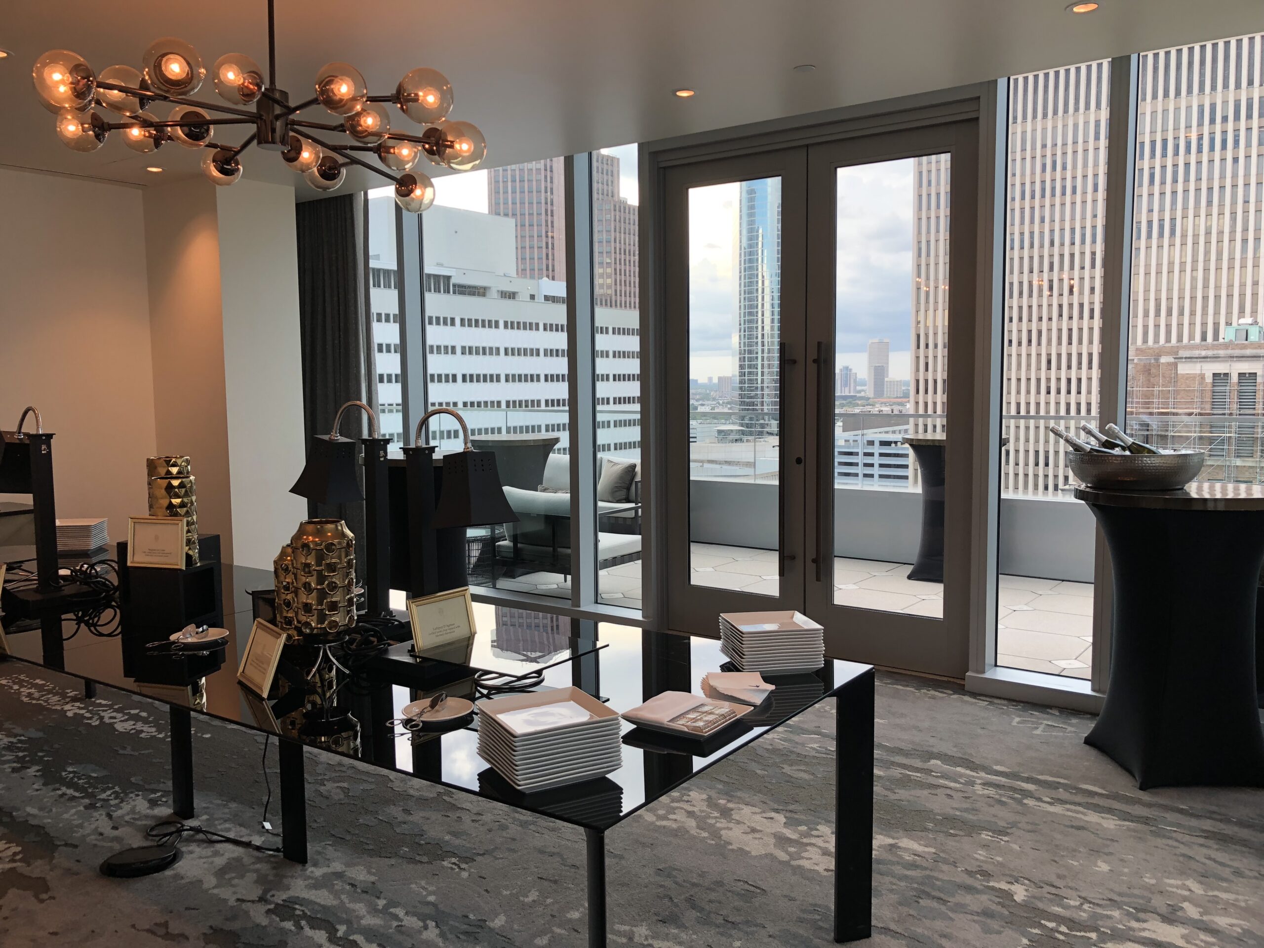 Modern conference room with glass doors opening to a city-view balcony, set with plates, bowls, and gift bags on a black table.