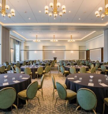 Spacious banquet hall with round tables, green chairs, black tablecloths, and chandeliers, set up for an event or conference.