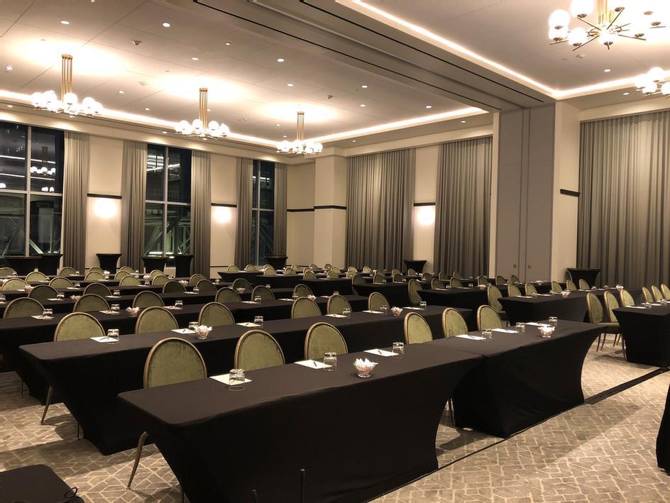 A large conference room with rows of tables and chairs, set up for a meeting. Each table has glasses, notepads, and bowls of candy.