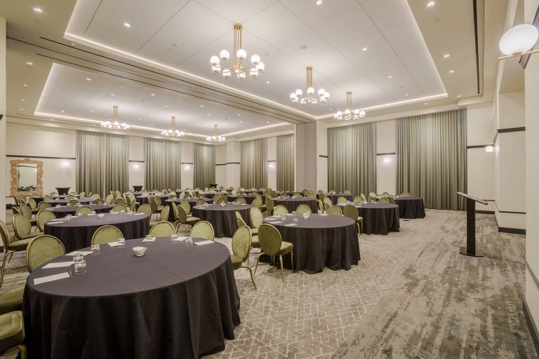 Spacious banquet hall with round tables, green chairs, chandeliers, and neutral décor, set up for an event or conference.