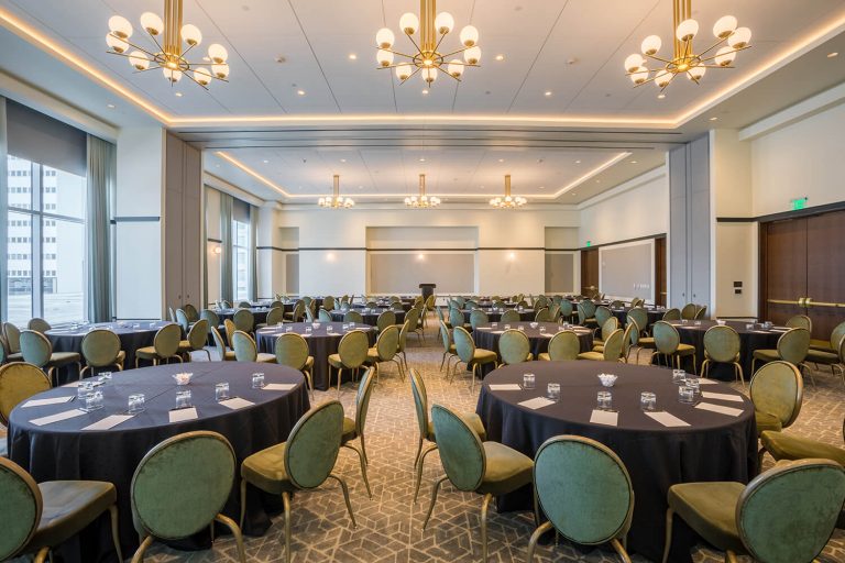 Spacious banquet hall with round tables, green chairs, black tablecloths, and chandeliers, set up for an event or conference.