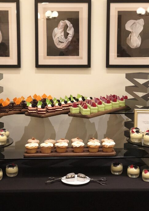 A dessert table with assorted cakes, cupcakes, and parfaits, arranged on black stands, with framed abstract art hanging on the wall behind.